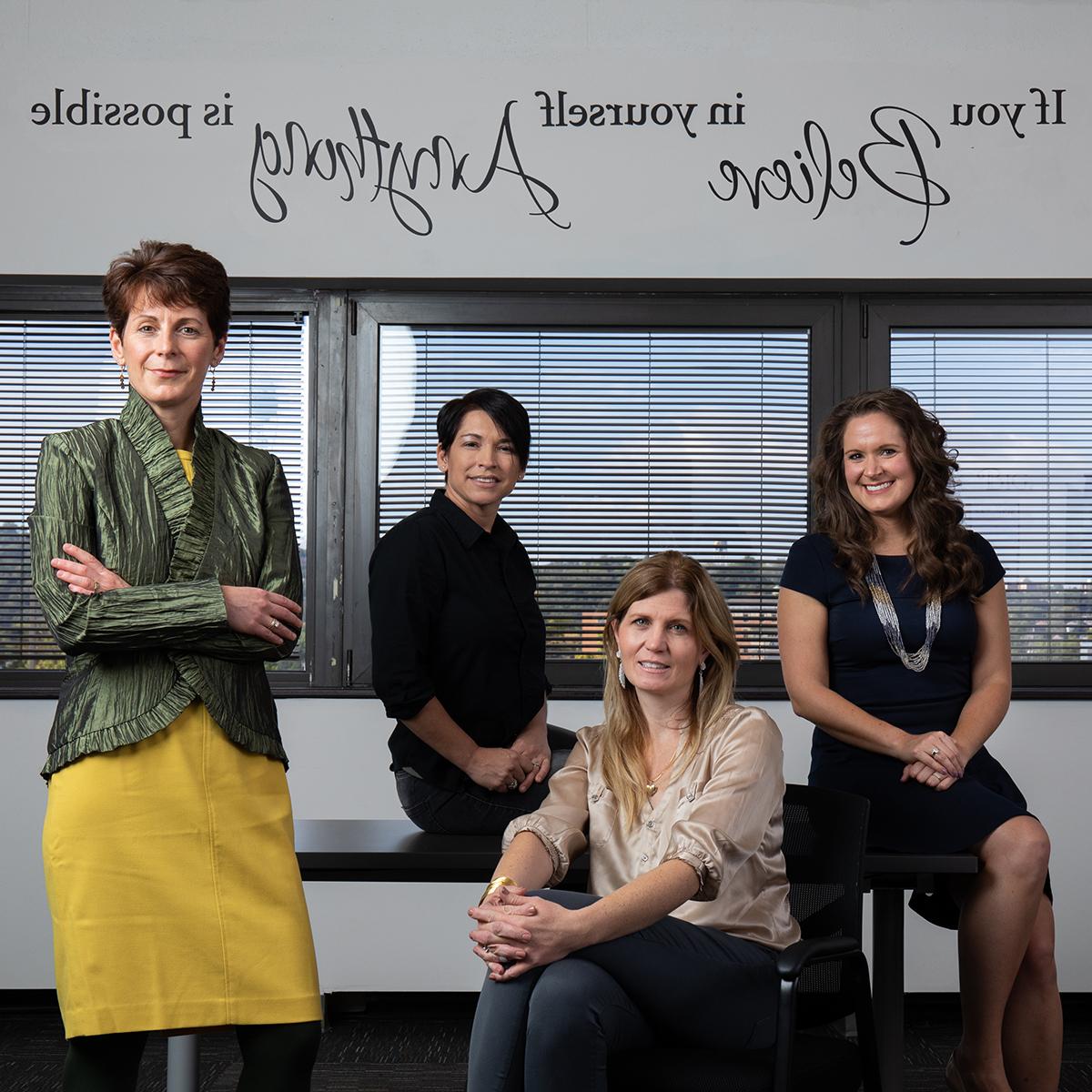 Photo of four women in professional clothing posing in front of a wall upon which is written the quote, "If you believe in yourself anything is possible"